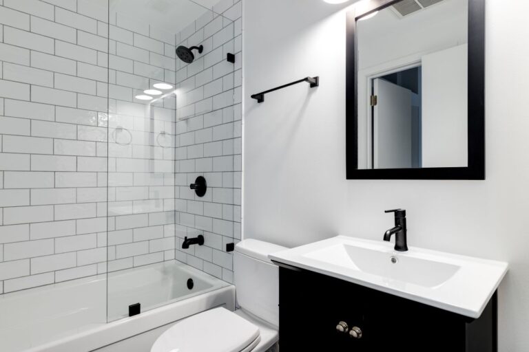A small modern bathroom with a dark vanity, mirror frame, and hardware. White subway tiles line the bathtub and shower with black faucets. New bathroom fixtures.
